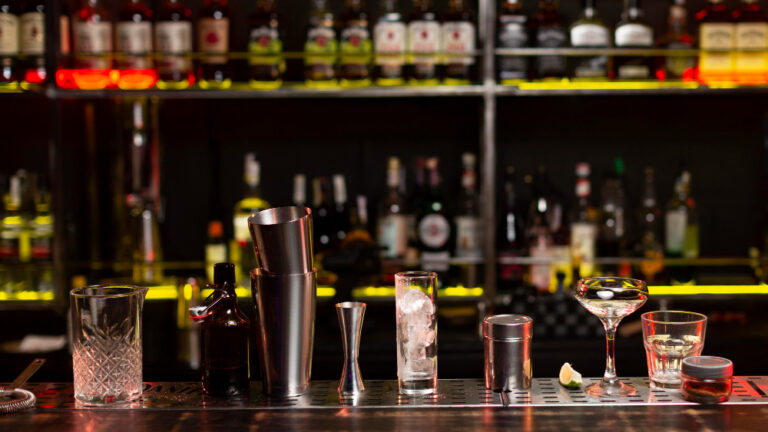 Eine beleuchtete Bar mit Flaschen im Hintergrund und verschiedenen Gläsern und Cocktail-Shakern auf der Theke.