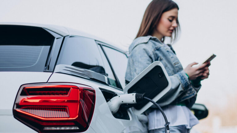 Eine junge Frau lehnt sich an ein weißes Auto, das gerade lädt, und schaut auf ihr Smartphone.