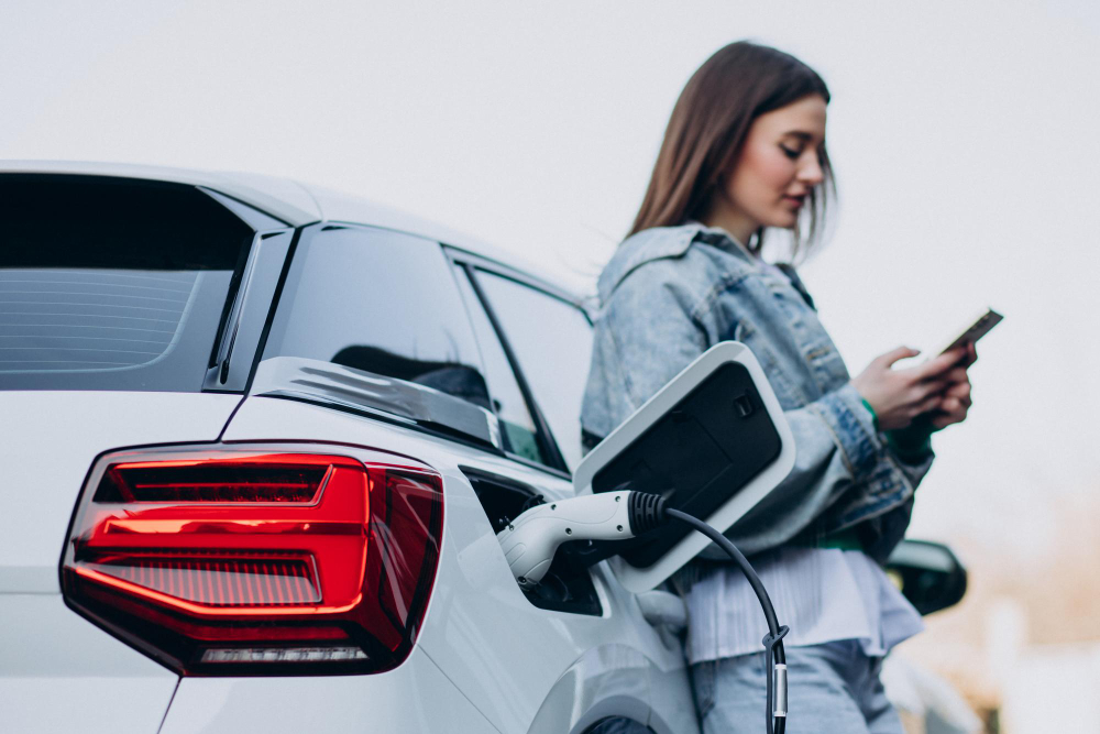 Eine junge Frau lehnt sich an ein weißes Auto, das gerade lädt, und schaut auf ihr Smartphone.