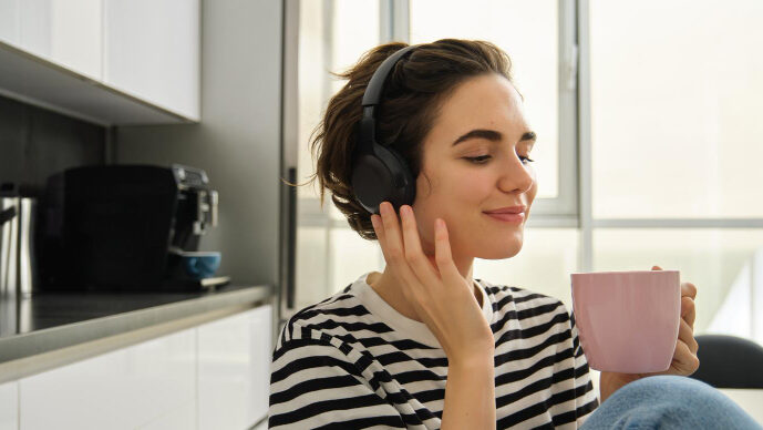 Eine lächelnde Frau in der Küche hört über Kopfhörer Musik und hält eine rosafarbene Tasse.