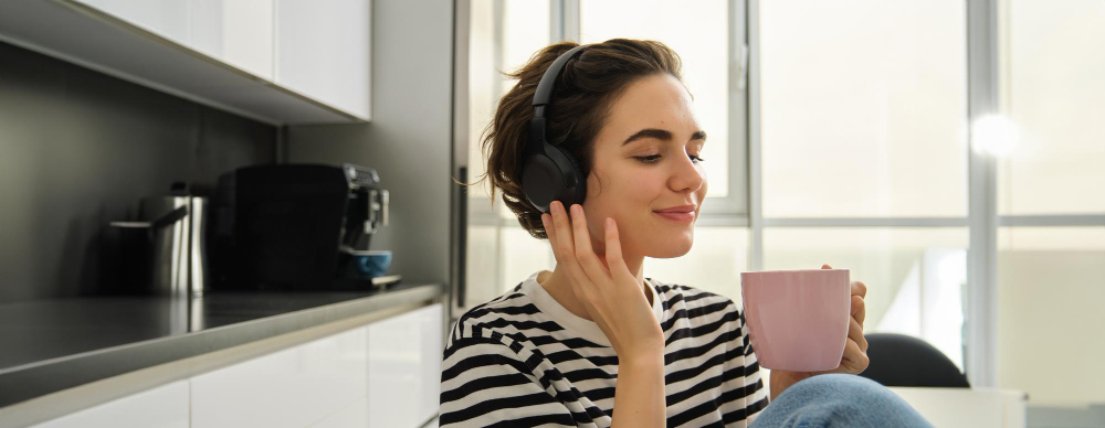 Eine lächelnde Frau in der Küche hört über Kopfhörer Musik und hält eine rosafarbene Tasse.