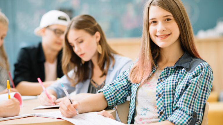 Eine lächelnde junge Frau schreibt in der Klasse, während sie in die Kamera schaut, und andere Schüler sitzen dahinter.