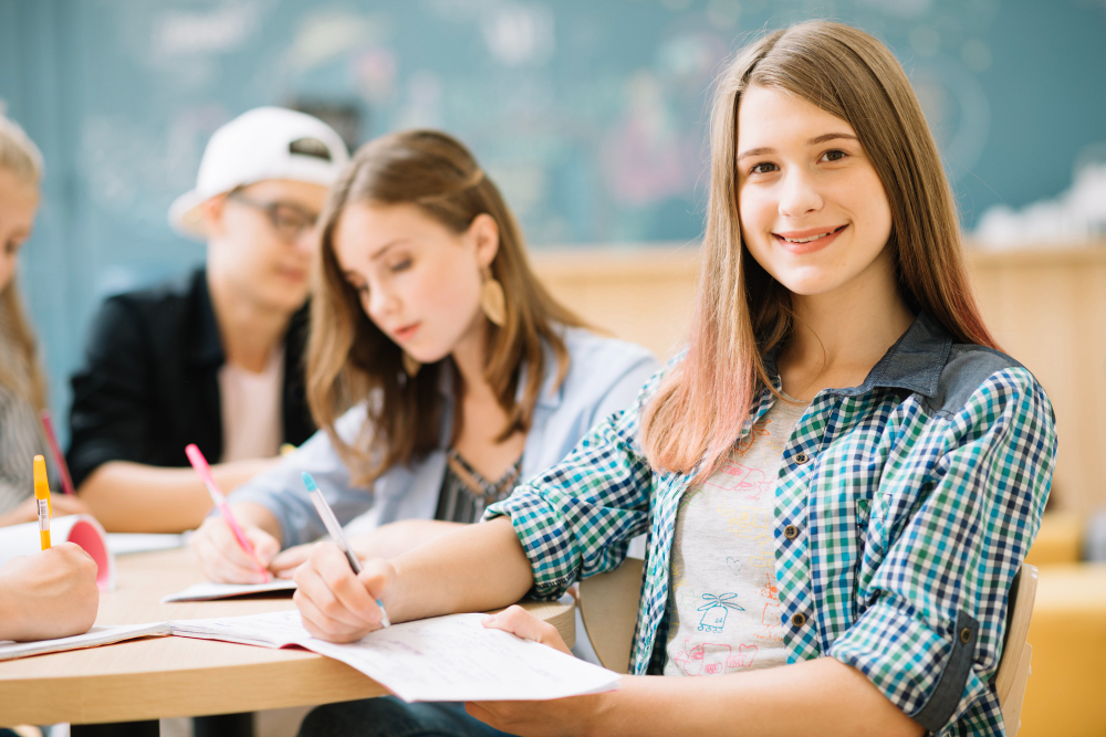 Eine lächelnde junge Frau schreibt in der Klasse, während sie in die Kamera schaut, und andere Schüler sitzen dahinter.