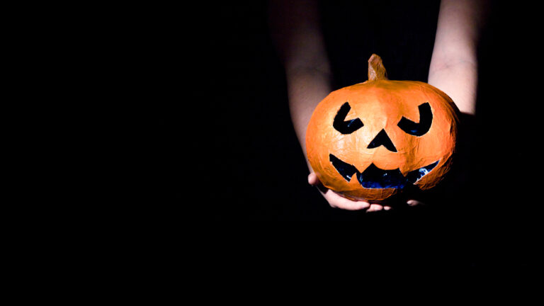 Zwei Hände halten eine orangefarbene Kürbis-Laterne (Jack O'Lantern) in einem dunklen Raum.