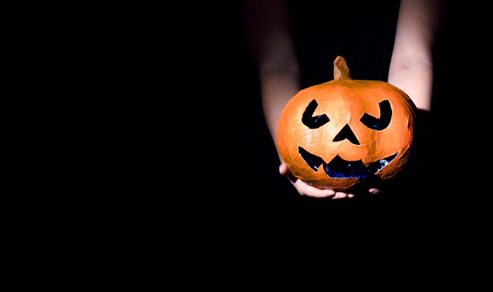 Zwei Hände halten eine orangefarbene Kürbis-Laterne (Jack O'Lantern) in einem dunklen Raum.