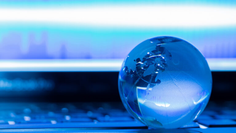 Eine kleine, blau beleuchtete Glaskugel, die die Erde darstellt, liegt auf einer Tastatur vor einem unscharfen Bildschirm.