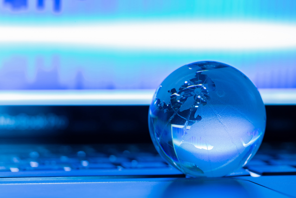 Eine kleine, blau beleuchtete Glaskugel, die die Erde darstellt, liegt auf einer Tastatur vor einem unscharfen Bildschirm.