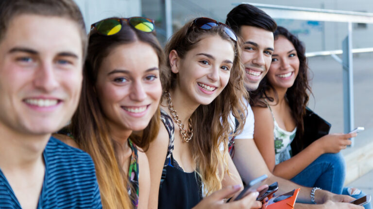 Fünf junge Erwachsene sitzen lächelnd zusammen und schauen auf ihre Smartphones.
