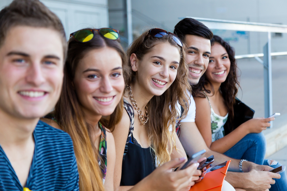 Fünf junge Erwachsene sitzen lächelnd zusammen und schauen auf ihre Smartphones.